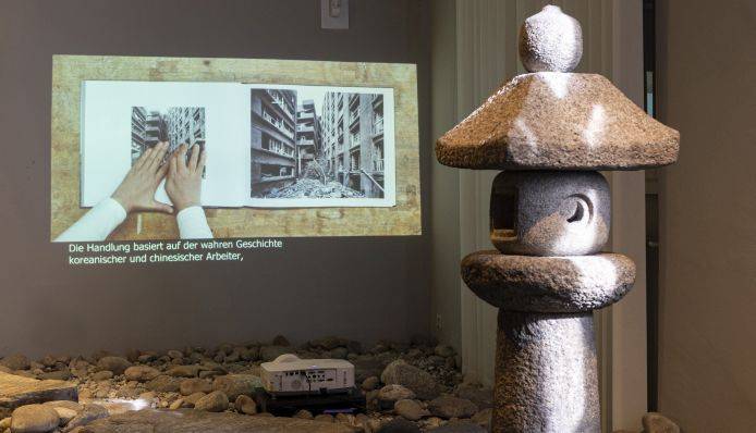 Videoprojektion im Steingarten der Ostasien-Dauerausstellung. Im Video wird ein Buch durchgeblättert, zwei Hände verdecken ausgewählte Bilder