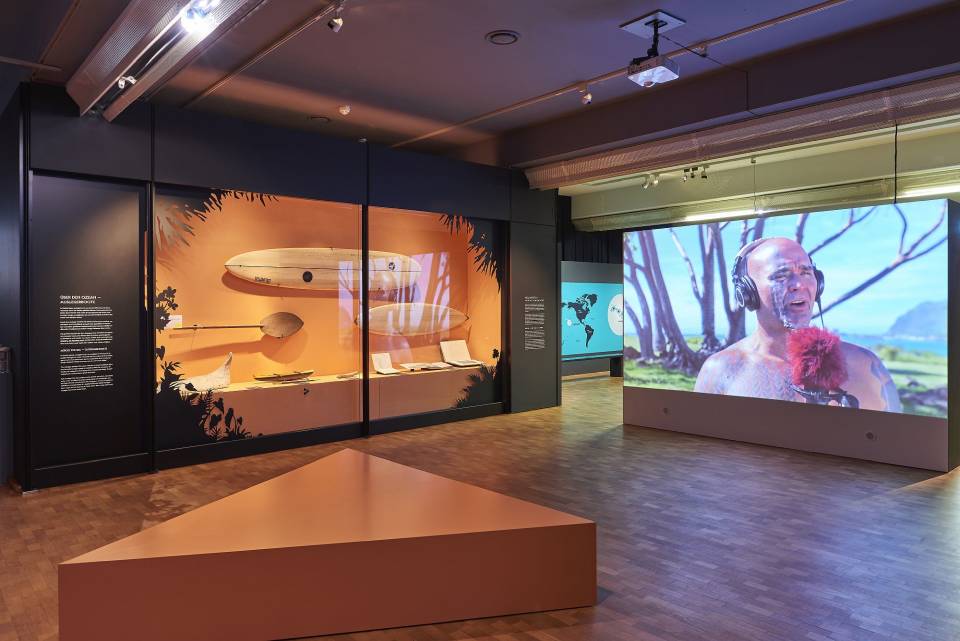 Ausstellungsansicht Hawaii. Rechts im Raum eine große Videoprojektion mit einem Interview, links eine Vitrine mit Surfbrettern und Paddeln