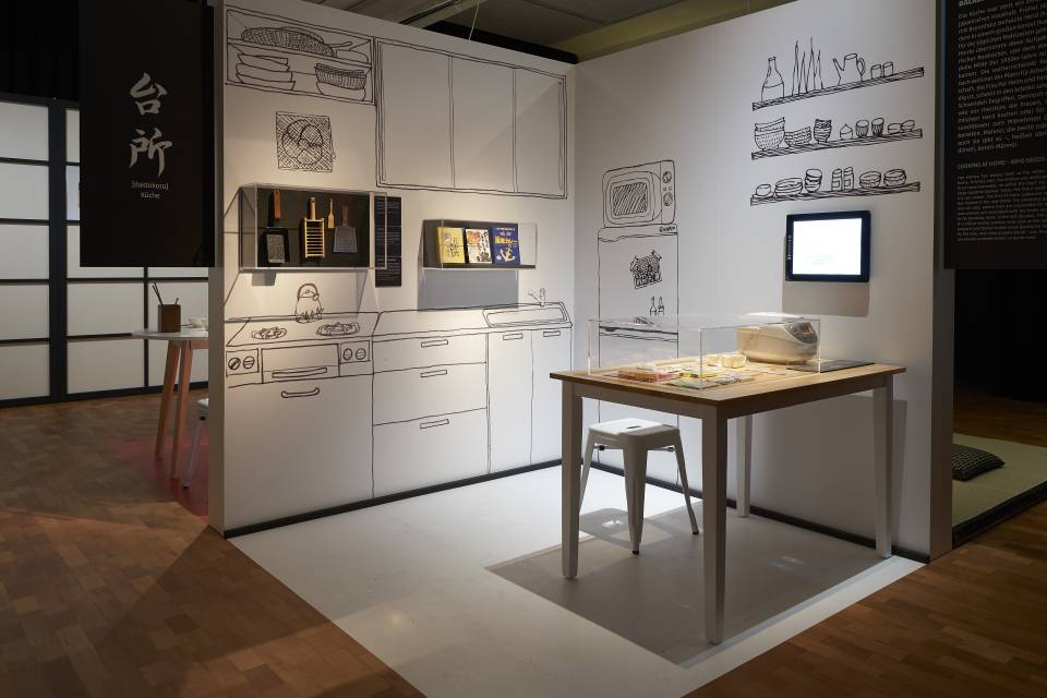 Ausstellungsansicht Oishii! Ein Esstisch mit Hocker steht vor einer an die Wand skizzierten Küche