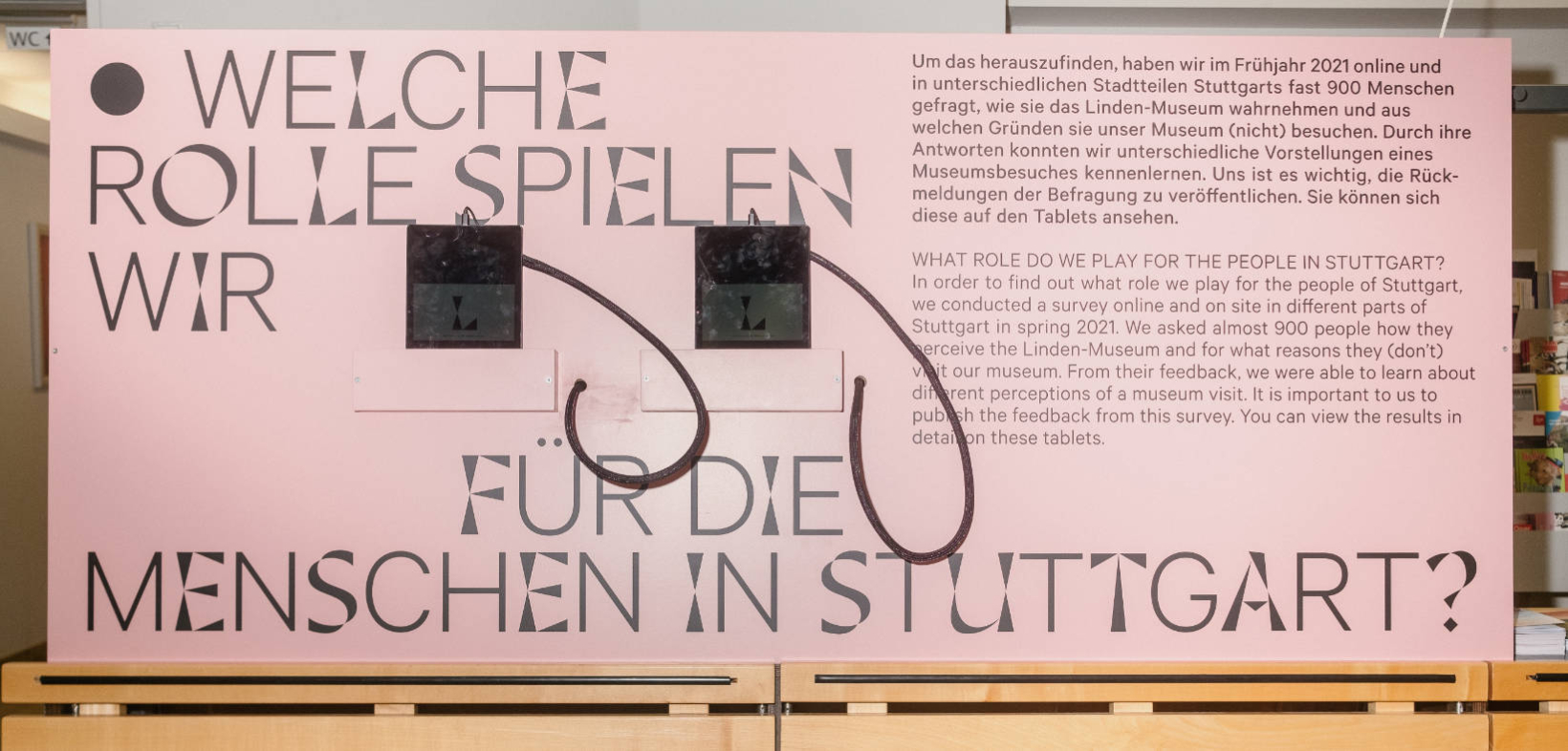 Präsentation LAB 8: Im Garderobenraum des Museums steht eine rosafarbene Tafel mit der Frage "Welche Rolle spielen wir für die Menschen in Stuttgart?", an der Tafel sind zwei Bildschirme befestigt
