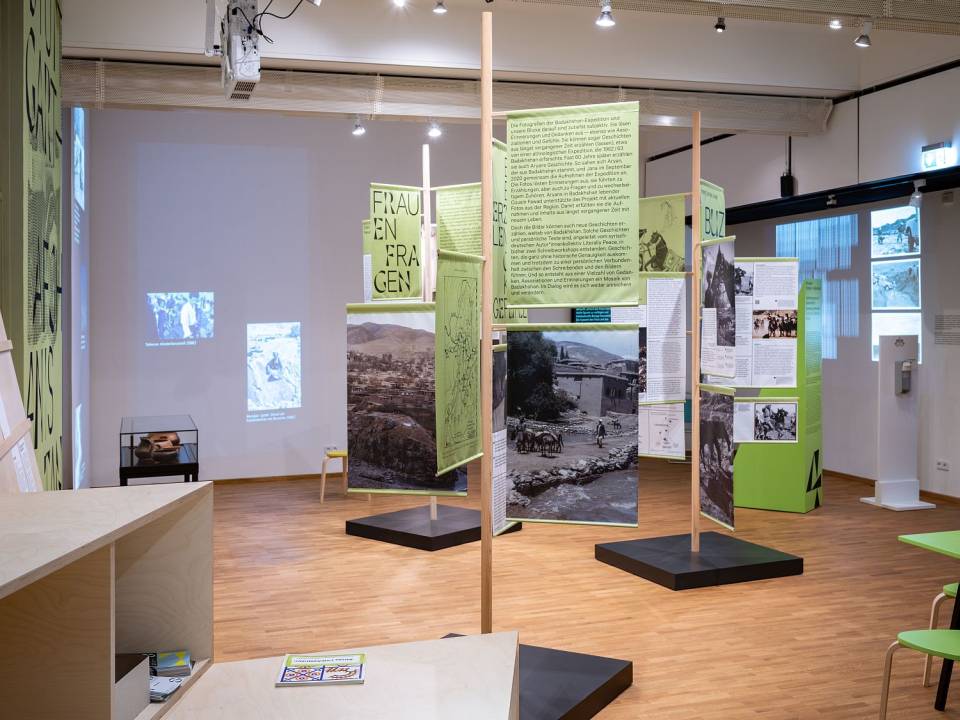 Präsentation von LAB 4: Im Ausstellungsraum stehen drei baumartige Konstruktionen mit Text- und Bildbannern daran. Im Hintergrund laufen zwei Projektionen