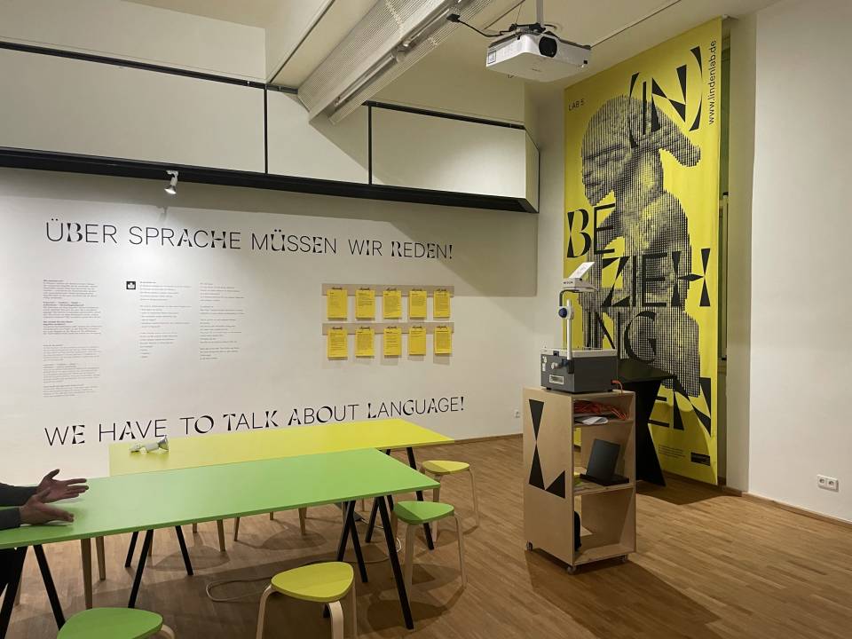 Präsentation von LAB 5: An einer Wand des LAB-Raums steht "Über Sprache müssen wir reden!", davor stehen zwei Arbeitstische. Rechts hängt ein gelbes Banner mit dem Keyvisual des Projekts