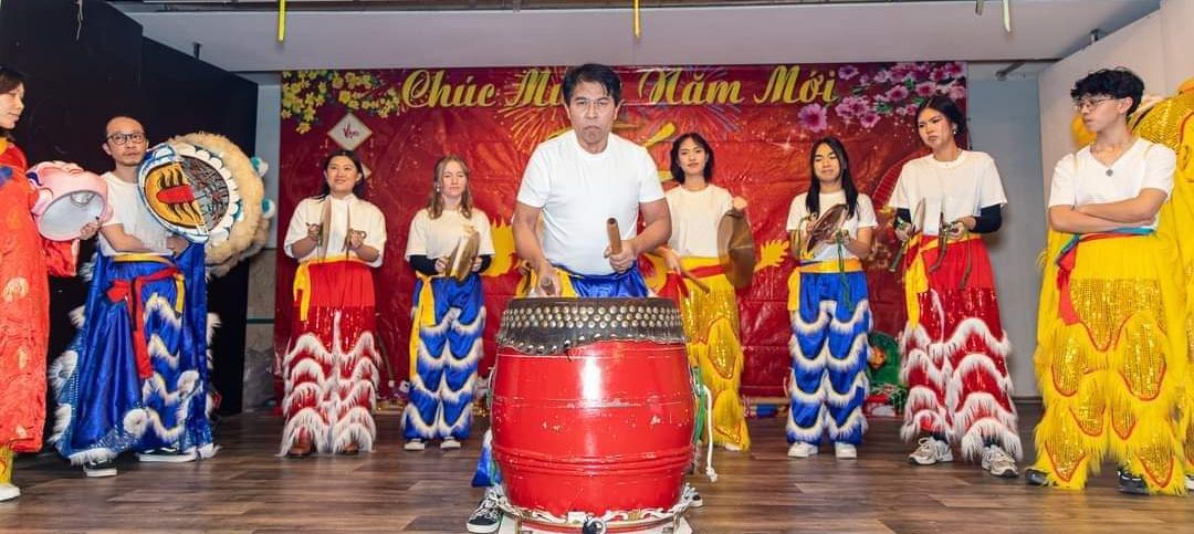 9-köpfige vietnamesische Musikgruppe mit langen Röcken in blau, rot und gelb. Im Vordergrund ein Trommler mit einer großen roten Trommel