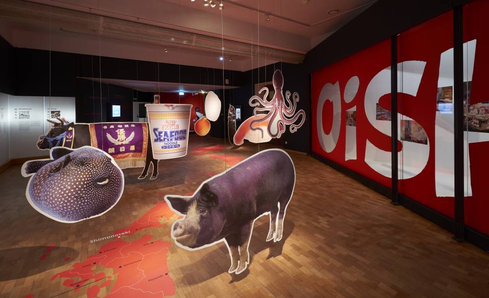 Ausstellungsansicht Oishii! Im ersten Ausstellungsraum hängen übergroßen Bilder von japanischen Lebensmitteln, wie Tintenfisch, Cup Noodles und ähnlichem von der Decke. Auf dem Boden ist eine Landkarte Japans abgebildet
