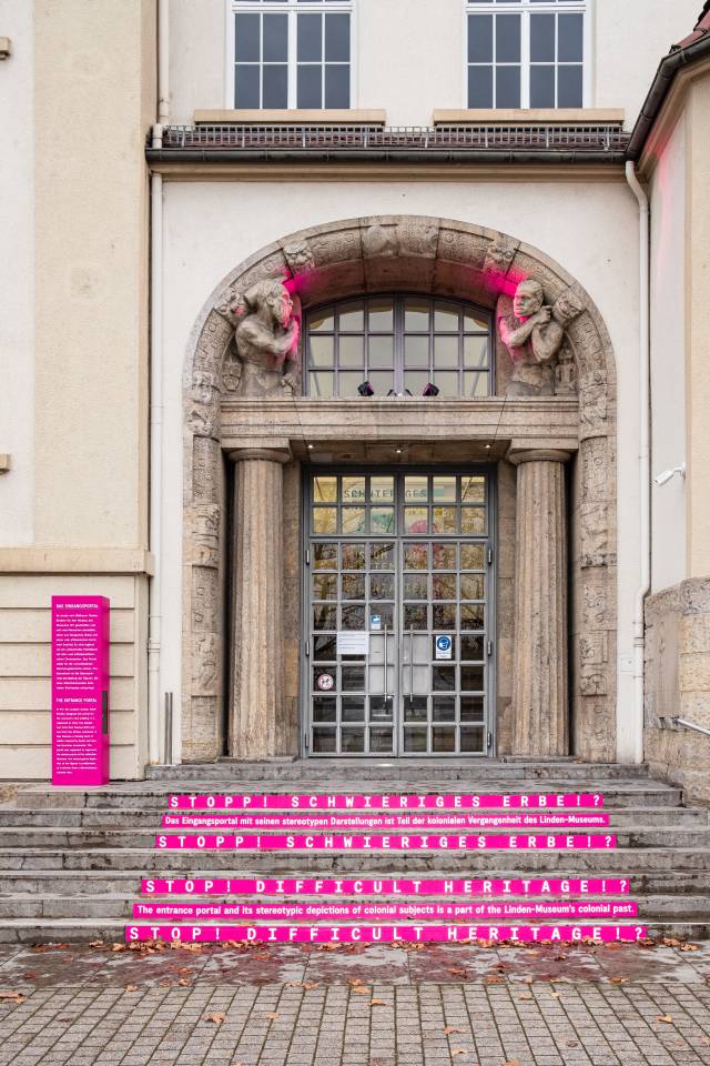 das Portal des Linden-Museums ist pink angestrahlt, die Treppenstufen mit pinken Streifen beklebt. Darauf steht "Stop! Schwieriges Erbe!?"