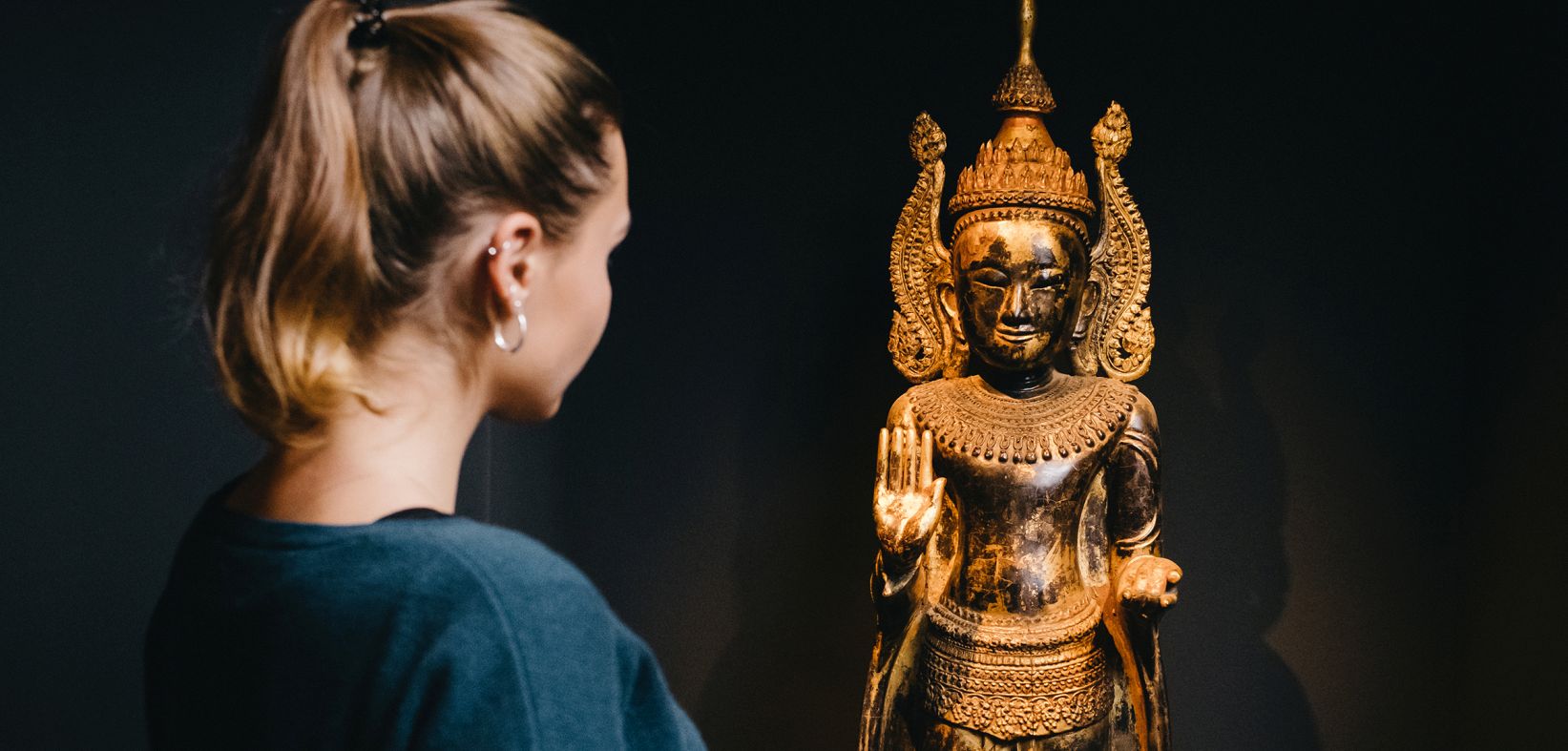 Junge Museumsbesucherin mit Pferdeschwanz blickt auf ein gekrönten Buddha aus dunklem Holz mit Gold überzogen.