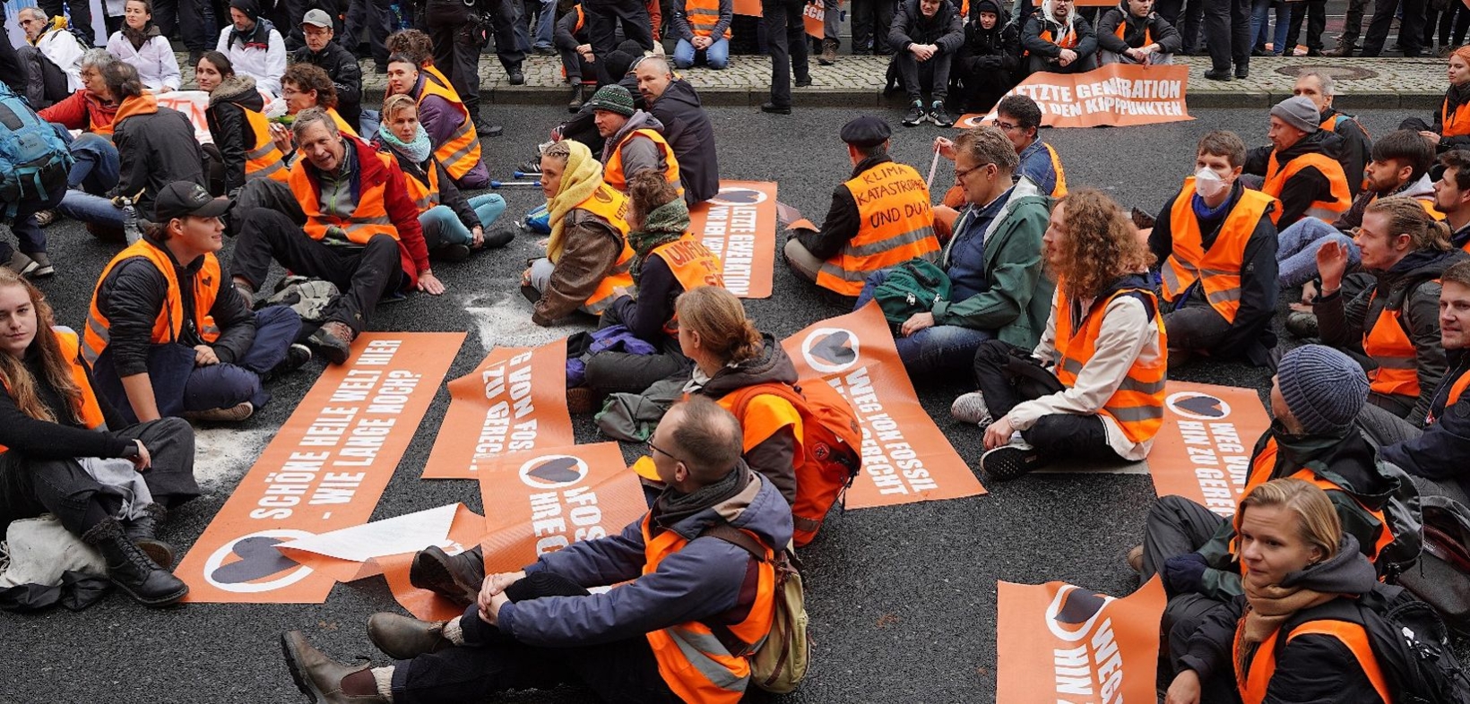 Straßensitzblockade der Letzten Generation: Rund 40 Demonstrant:innen, vornehmlich mit orangen Westen und orangen Transparenten auf der Straße, am Straßenrand Polizist:innen