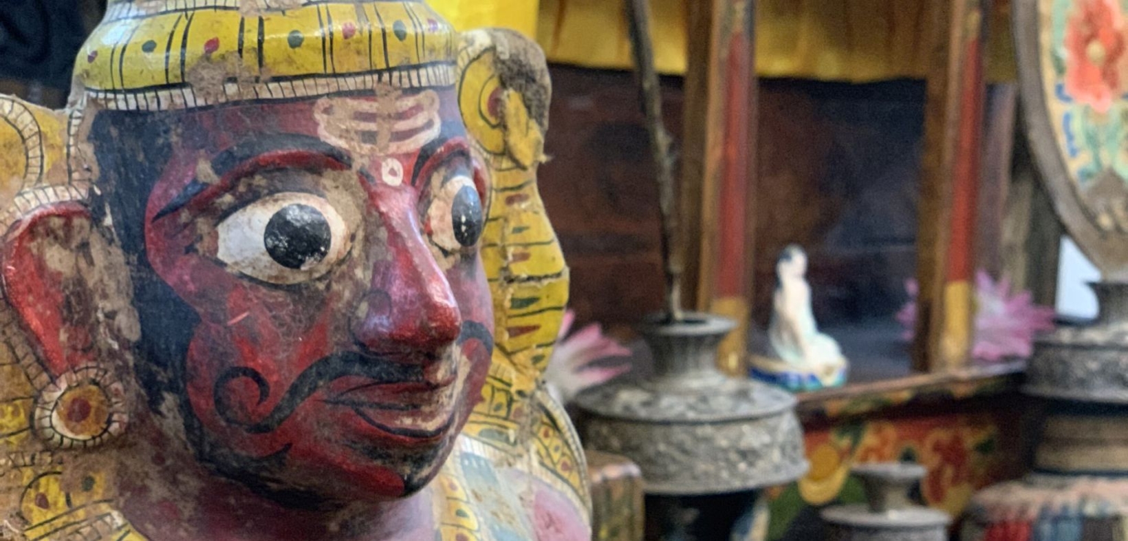 Große Keramikstatue mit rotem Gesicht, gelbem Kopfschmuck und großen Augen , im Hintergrund ein Markt in Nepal