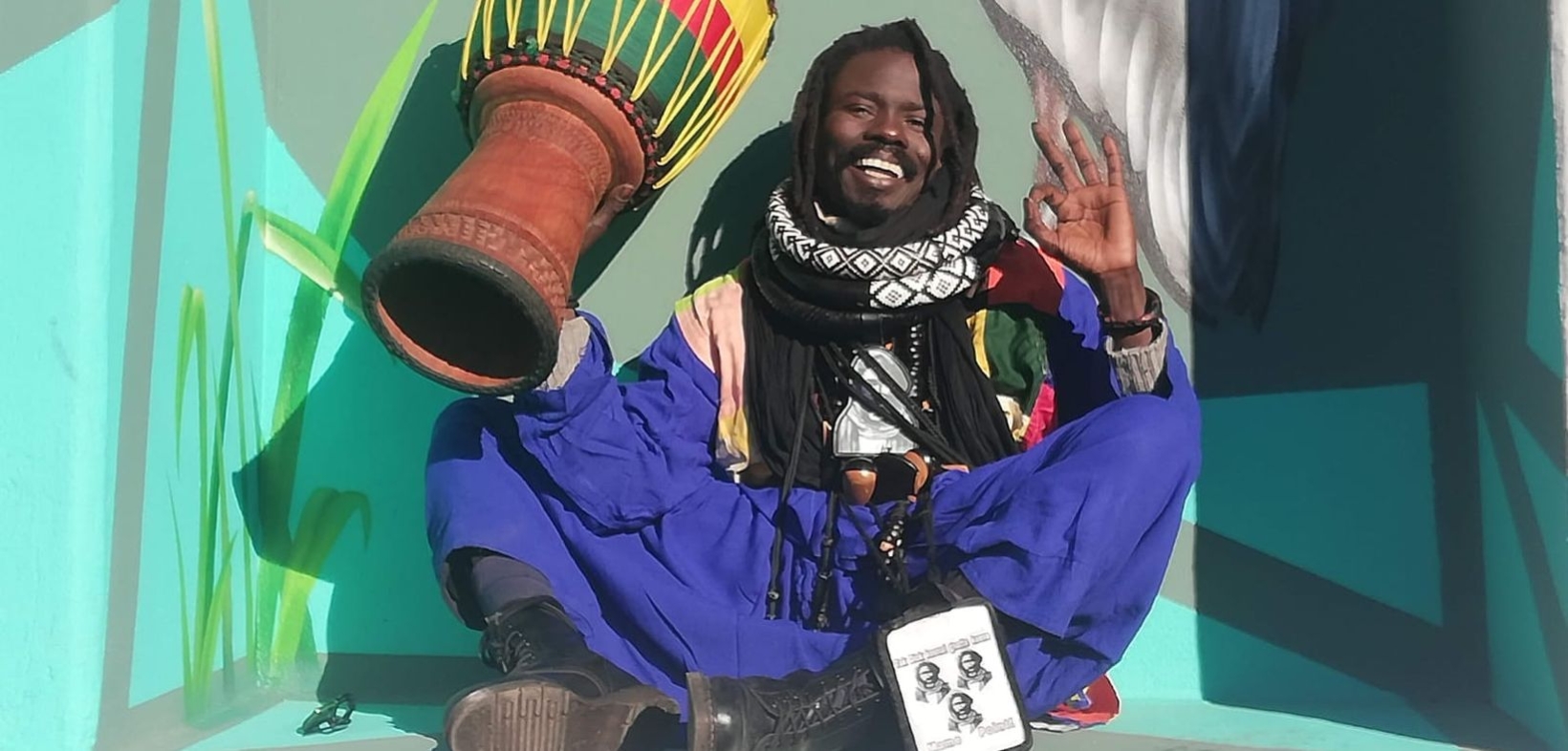 Mame Cheikh Seck: Schwarzer Mann mit Dreadlocks, lachend mit erhobenen Händen; in der einen Hand hält er eine Djembe in die Luft, mit der anderen formt er das "Alles bestens"-Zeichen; er hockt auf dem Boden und trägt einen schwarzen Pulli mit bunten Applikationen und eine weite blaue Hose