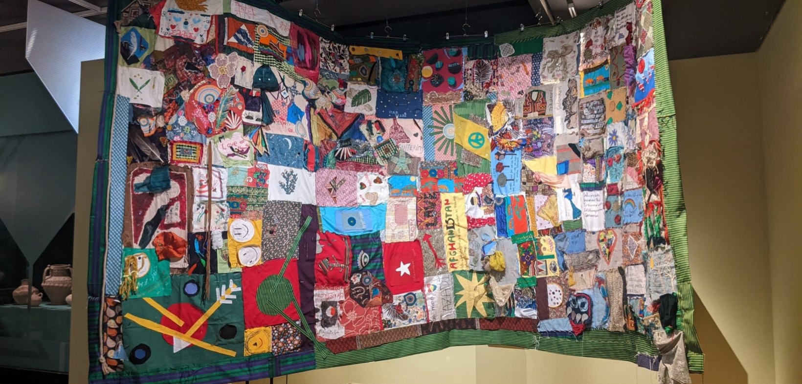 Patchwork-Flagge aus vielen kleinen zusammengenähten Stoffresten. Ein Projekt des Künstlers Aman Mojadidi, in einem partizipativen Prozess mit Mitgliedern der Projektgruppe "Entangled: Stuttgart - Afghanistan" und Besucher:innen entstanden. Mehrere Meter breit und hoch im Raum hängend.