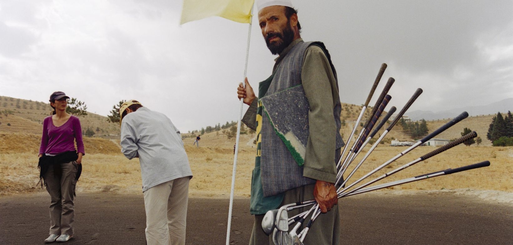 Relativ ausgetrockneter Golfplatz mit zwei weißen Golfspieler:innen im Hintergrund, im Vordergrund ein afghanischer Mann als Assistent mit vielen Golfschlägern und Fahne für das Golfloch