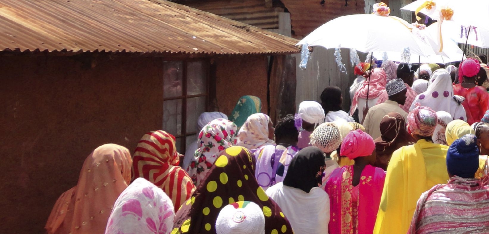 Prozession im Slum Kib(e)ra in Nairobi: Draufsicht von hinten auf buntgekleidete Menschen, alle mit Kopftuch oder Käppi; im Zentrum der Prozession zwei weiße geschmückte Schirme