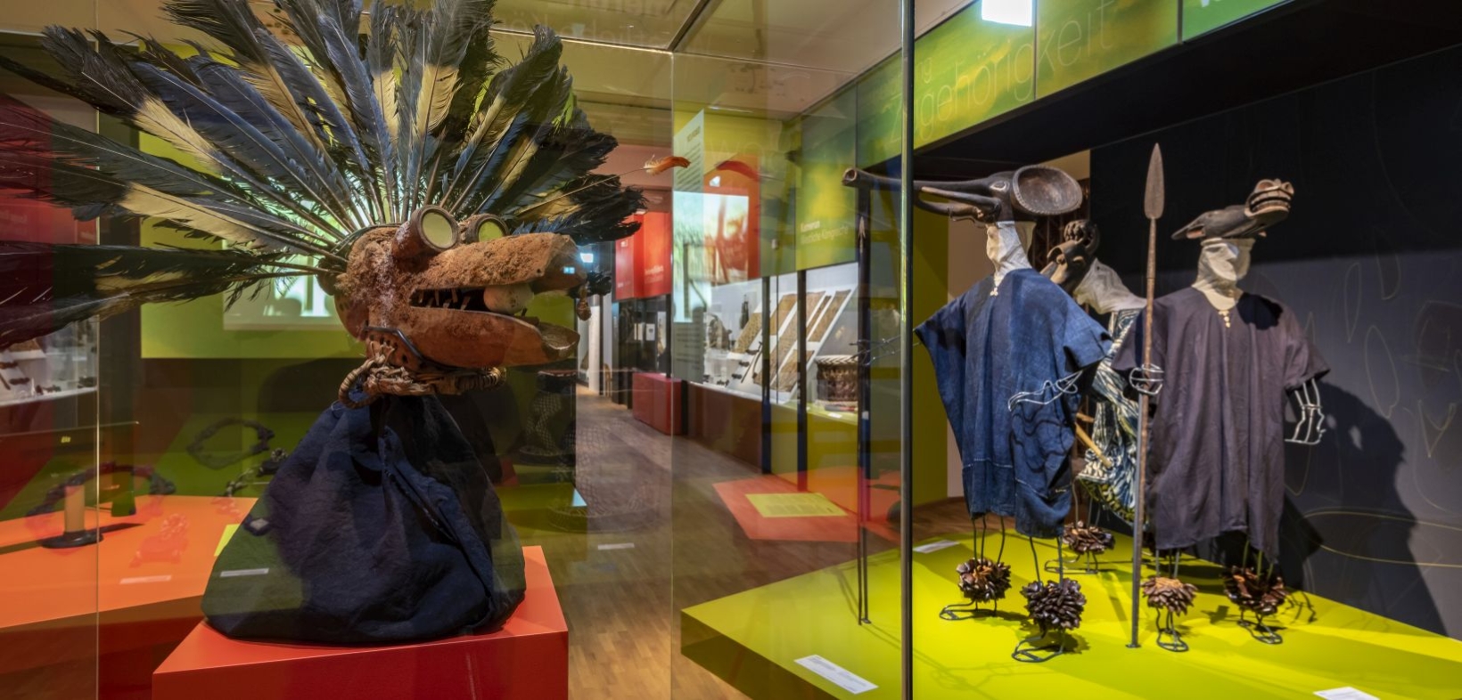 Blick in die Ausstellung "Wo ist Afrika?"; im Vordergrund eine Maske mit Federn, einer Spiegelbrille und einem Ei in einer langen Schnauze; im Hintergrund eine Maskenzug aus Kamerun