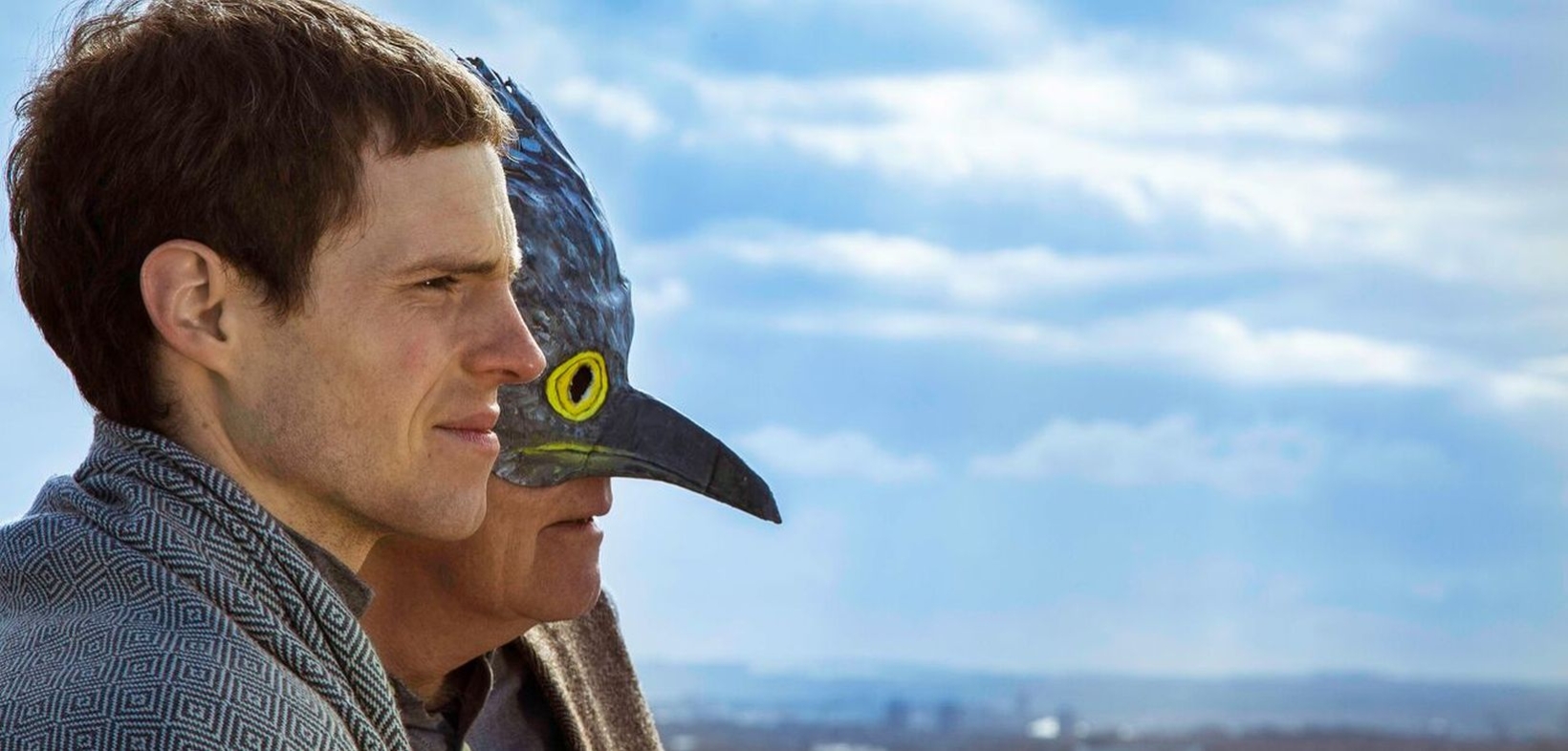 Weiter strahlender Himmel mit weißen Wolken. Vom linken Bildrand blicken Joshua Green und (verdeckt) sein Vater Malcolm Green mit Kuckucksmaske im Profil in die offene Landschaft