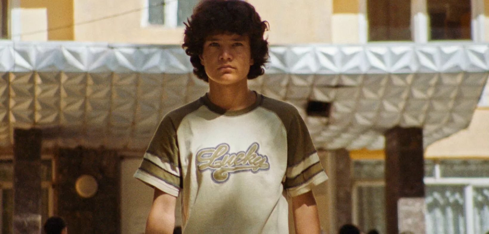 Lockiger Junge im braun-weißen T-Shirt mit der Aufschrift "Lucky" vor Gebäude
