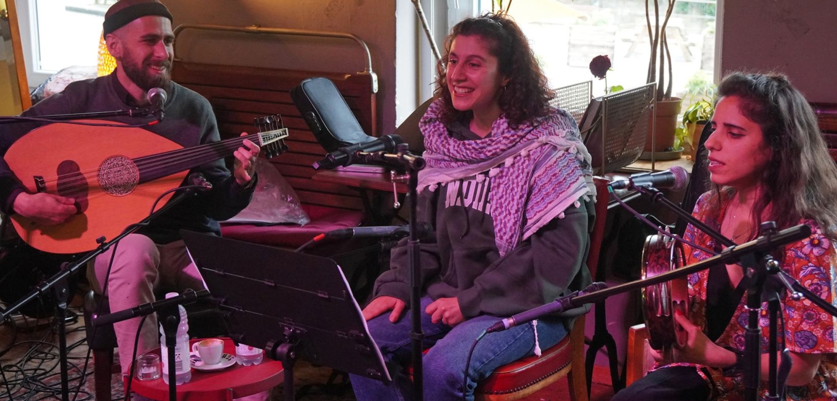 von links nach rechts: ein männlicher Musiker mit Oud, eine Sängerin und eine Musikerin mit Tamburin spielen zusammen in einem Café