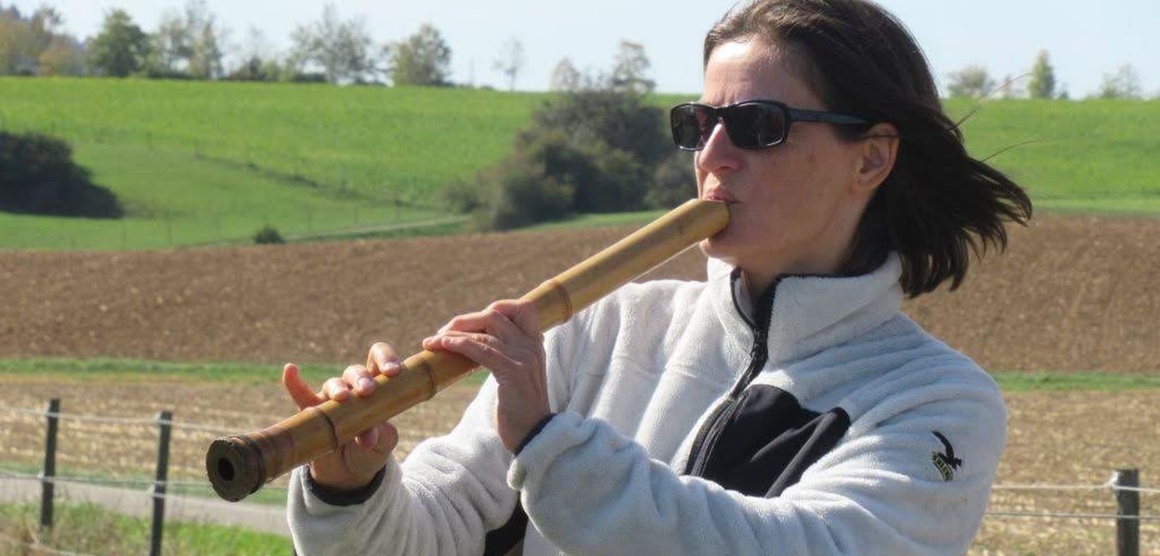 Sylvia Shoin-Bittmann (Sonnenbrille, dunkle mittellange Haare, weiße Fleecejacke) spielt im Freien Shakuhachi, im Hintergrund hügeliges Ackerland