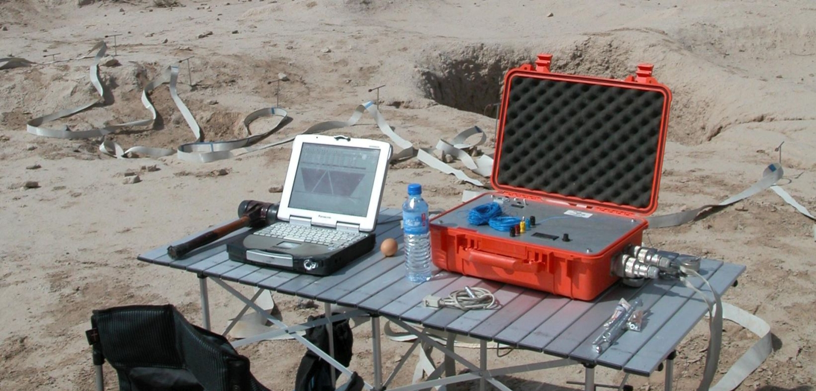 silberner Klapptisch mit Laptop, rotem Koffer und Wasserflasche, einem schwarzen Klappstuhl davor, an Grabungsstätte in wüstenartigem Gebiet