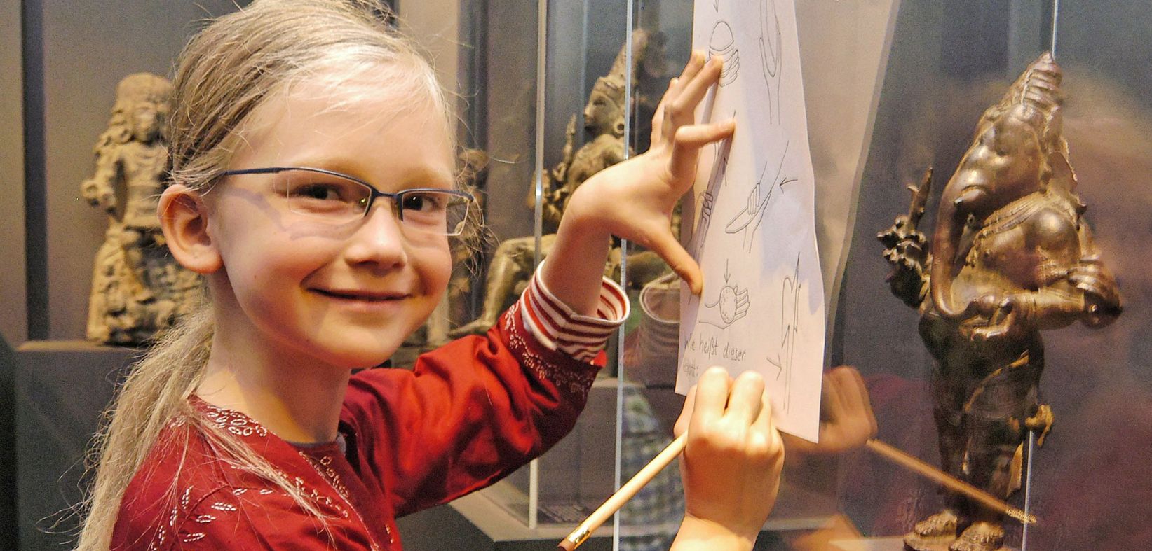 Ein kleines blondes Mädchen zeichnet auf einem Blatt welches sie auf der Fensterscheibe der Vitrine abstützt. In der Vitrine ist eine goldene Elefantenfigur zu sehen.