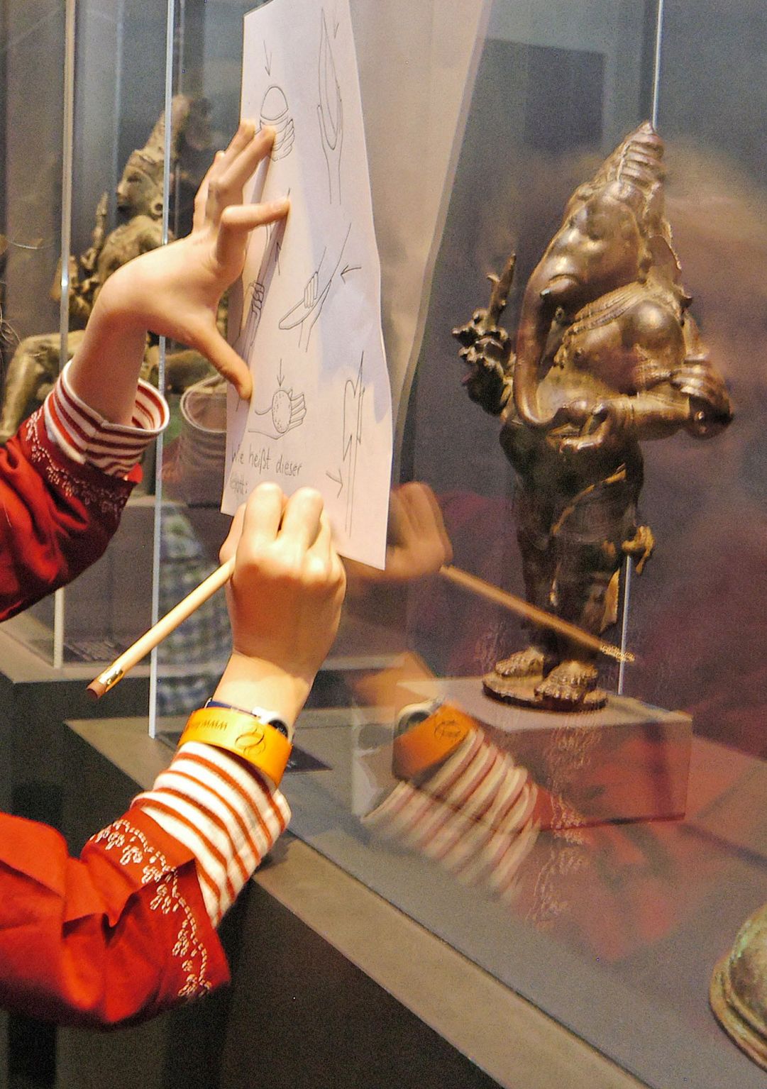 Ein kleines blondes Mädchen zeichnet auf einem Blatt welches sie auf der Fensterscheibe der Vitrine abstützt. In der Vitrine ist eine goldene Elefantenfigur zu sehen.