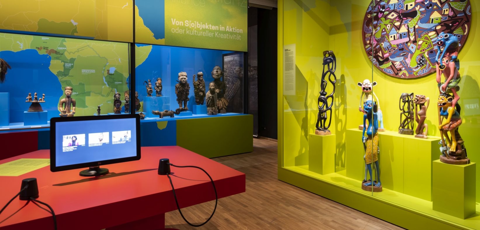 Blick in die Ausstellung "Wo ist Afrika?". Im Vordergrund Medienstation auf rotem Tisch, im Hintergrund Skulpturen in blau und grün gestalteten Vitrinen