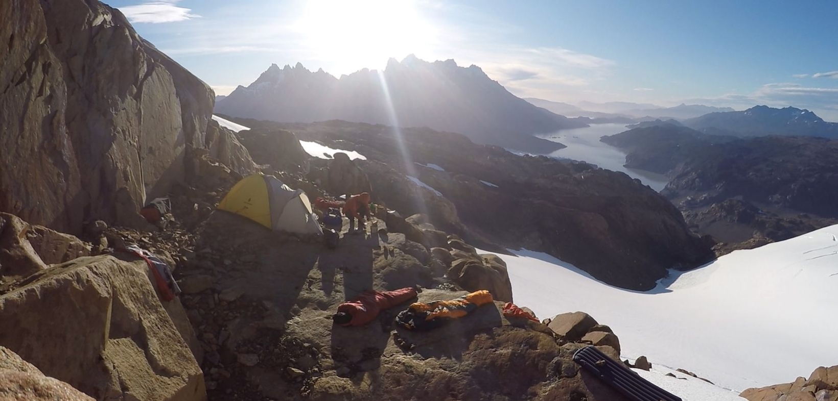 Sonnenbeschiene Gebirgslandschaft am Morgen; auf einem Vorsprung ein Biwaklager, Blick auf Schneefelder und einen See