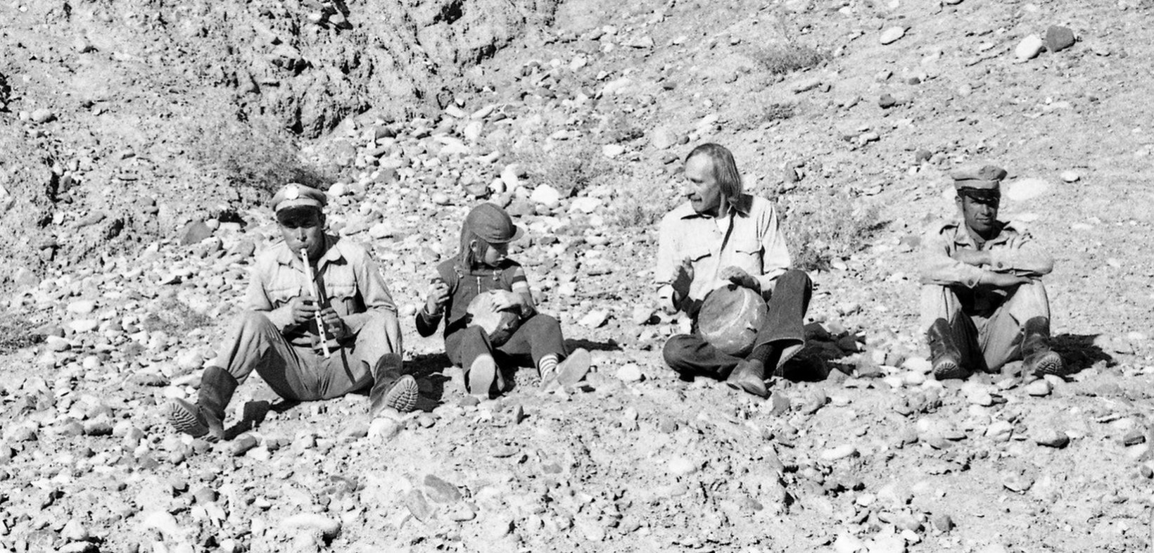 Hartmut Geerken musiziert sitzend auf Geröll mit zwei anderen afghanischen Männern. Einer spielt Flöte, zwei spielen Tabla, einer vierter Mann sitzt rechts daneben ohne Instrument. Alle sitzen auf Geröll in einer felsigen Landschaft. Schwarzweiß-Aufnahme