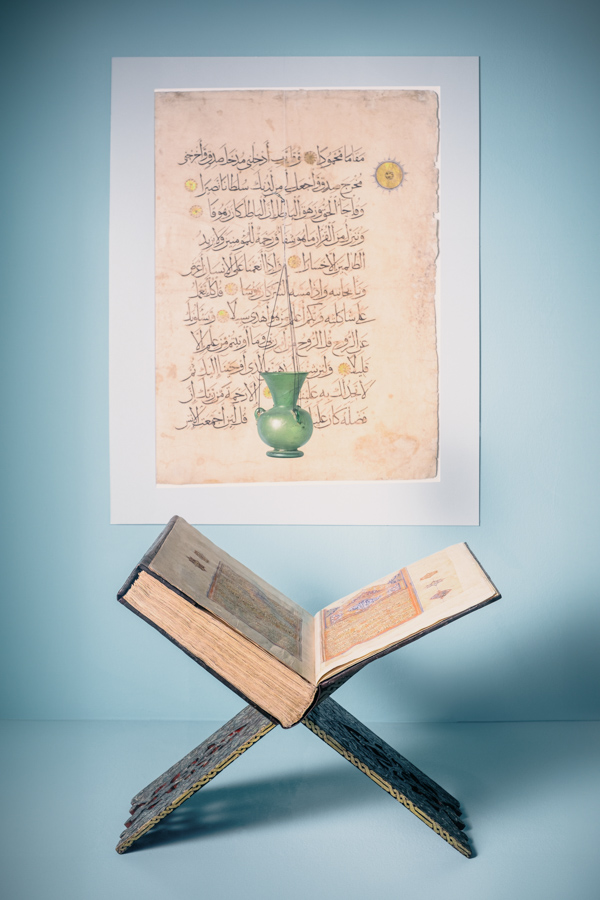 Ein Blatt aus einem Koranmanuskript für eine Moschee hängt an einer türkisfarbenen Wand. Davor steht ein aus Holz geschnitzter klappbarer Koranständer (kursi) mit einem Prachtkoran. Darüber hängt eine grüne Moscheelampe aus Glas