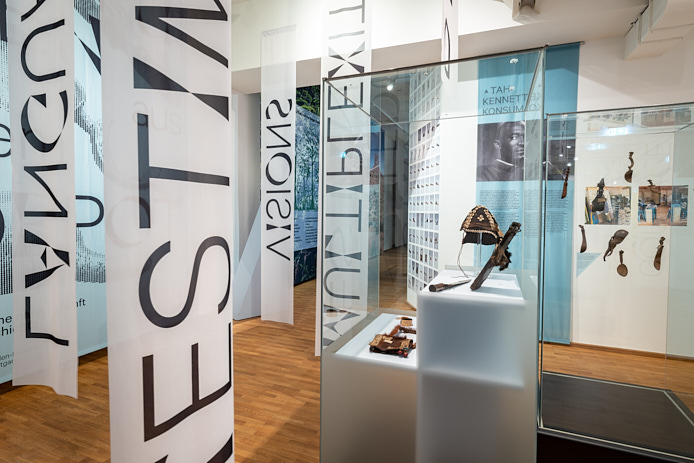 Blick in den Ausstellungsraum von LAB 6. Links hängen Banner mit Text von der Decke, rechts stehen Vitrinen mit Objekten aus der Sammlung Bertram